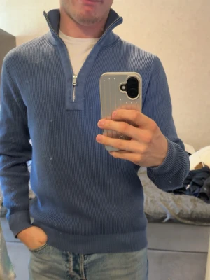 Blå ribbstickad tröja med halvzip från j lindeberg - Snygg blå ribbstickad tröja med halvzip och hög krage. Tröjan har långa ärmar och breda muddar vid ärmslut och nederkant. Perfekt för dig som gillar en stilren och avslappnad look. Passar medium