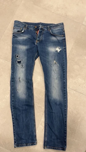 Blå  dsquared2 jeans med raka ben - Säljer ett par blå jeans med raka ben och slitningar på både fram- och baksida. Jeansen har klassisk femficksmodell, knappgylf och ljusa tvättade partier. Perfekta för en avslappnad och trendig look.