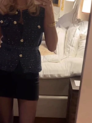 Svart glittrig blus med guldknappar - Snygg svart blus med glittriga detaljer och dekorativa guldknappar framtill. Modellen är ärmlös och har markerad midja med bälte. Perfekt för dig som vill sticka ut lite extra. Aldrig använd 