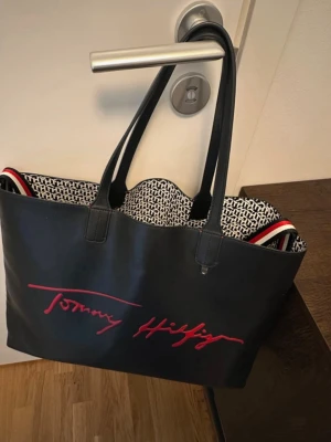 Svart shoppingväska Tommy Hilfiger - Rymlig väska från Tommy Hilfiger med avklippt detalj på framsidan som syns på tredje bilden. Den har även fått märken i kanterna som syns på fjärde bilden. Väskan i sig är knappt använd och därför inga tecken på användning i handtag eller liknande🌸