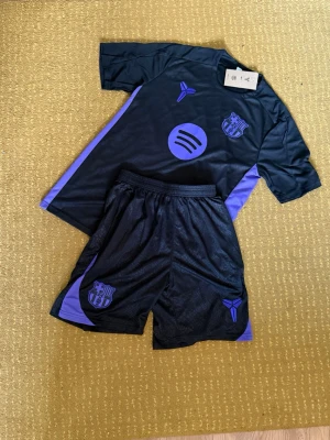  Barcelona träningsset - Säljer ett svart och lila träningsset från Barcelona med matchande t-shirt och shorts. Setet har lila detaljer längs sidorna och klubbmärke på både tröja och shorts. Materialet är lätt och andas, perfekt för träning eller fotboll. Snygg och sportig design.