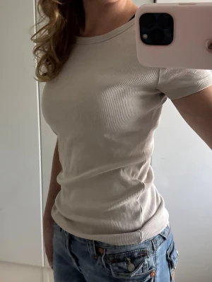 Beige ribbad t-shirt - Snygg beige ribbad t-shirt med rund hals och korta ärmar. T-shirten har en tight passform och sitter skönt mot kroppen. Perfekt basplagg att matcha med jeans eller kjol för en enkel och stilren look.
