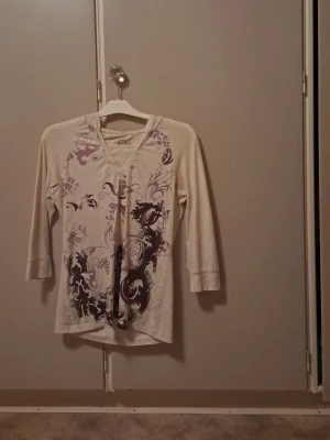 Ljuslila topp med mönster Style&co - Snygg unik vit topp från Style&co med v-ringning och trekvartsärmar. Toppen har ett unikt lila och mörklila blommigt mönster framtill som ger en cool vibe. Tillverkad i mjukt bomullsmaterial som känns skönt mot huden. 2000s & early 2010s vibe. Snygg med mörka jeans.