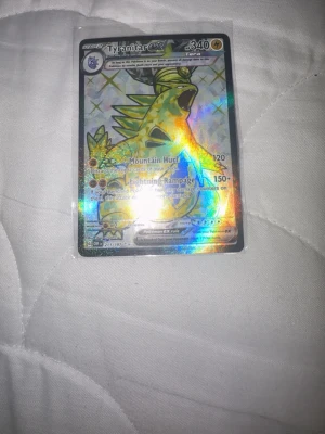 Tyranitar EX Pokémonkort holo - Säljer ett holografiskt Tyranitar EX Pokémonkort med coolt mönster och glansig yta. Kortet har färger i grönt, gult och svart och är inplastat för extra skydd. Perfekt för samlare eller Pokémonfans som vill utöka sin samling.
