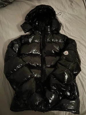 Svart glansig dunjacka från Moncler - Säljer en svart, glansig dunjacka från Moncler med huva och klassisk logga på ärmen. Jackan har en puffig design och dragkedja framtill. Perfekt för kalla vinterdagar och riktigt snygg streetstil.