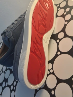 Gråa Louboutin sneakers med röd sula - Snygga grå sneakers från Christian Louboutin med klassisk röd sula och vita kanter. Skorna har en stilren design i mocka med matchande grå skosnören och diskreta sömmar. Perfekta för dig som vill sticka ut med exklusiva detaljer och ikonisk look.
