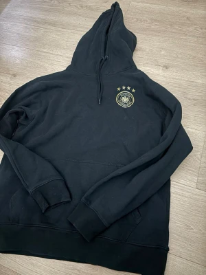 Svart hoodie Tyskland landslag - Svart hoodie med Tysklands landslagslogga och fyra stjärnor på bröstet. Stor tryckt text 'GER' och årtal på ryggen när de vanm VM, samt guldstjärnor. Klassisk känguruficka och dragsko i huvan. Perfekt för dig som älskar fotboll. 