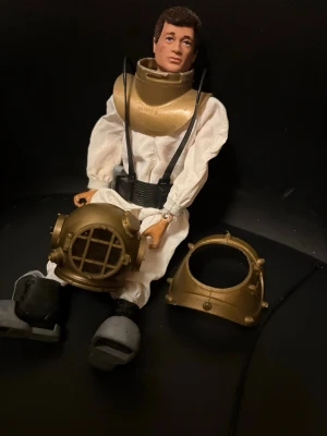Vintage dykardocka med hjälm - Säljer en cool vintage dykardocka med vit overall, svarta detaljer och avtagbar hjälm i guldigt plastmaterial. Dockan har realistiska detaljer som viktbälte och stövlar. Perfekt för samlare eller som retropryl till hyllan. Från 1964 England 