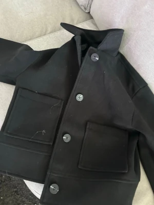 Svart overshirt med stora fickor - Stilren svart overshirt med raka linjer och stora utanpåliggande fickor framtill. Jackan har klassisk krage och stängs med fyra svarta knappar. Materialet ser ut att vara kraftigt tyg, perfekt för lager-på-lager. Enkel och clean design som passar till det mesta.