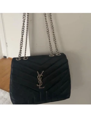 Svart YSL axelväska med kedja - Snygg svart axelväska från YSL med quiltat mönster och silverfärgad kedjerem. Väskan har klassisk YSL-logga i metall framtill och en stilren, rektangulär form. Perfekt för dig som gillar lyxiga accessoarer och tidlös design.