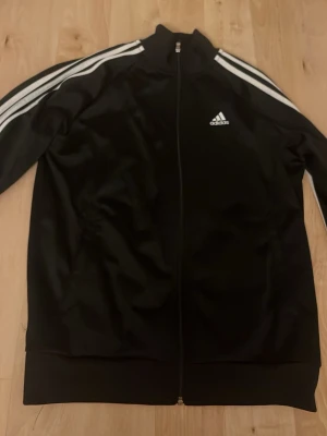 Svart Adidas träningsjacka - Klassisk svart träningsjacka från Adidas med vita ränder längs ärmarna och dragkedja framtill. Jackan har hög krage och Adidas-logga på bröstet. Tillverkad i ett glansigt syntetmaterial som är bekvämt och sportigt.