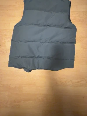 Svart dunväst med quiltad design - Stilren svart dunväst med quiltad look och breda horisontella sömmar. Västen har hög krage och är lätt vadderad för extra värme. Passar perfekt för lager-på-lager och har en klassisk, tidlös form. Märke och storlek syns på insidan. Xs men lite stor i storleken 