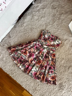 Blommig axelbandslös kortklänning - Superfin axelbandslös klänning med blommigt mönster i rosa, orange och gröna toner. Klänningen har en utställd kjol och markerad midja, perfekt för sommarens alla tillfällen. Materialet känns lätt och luftigt. Obs söm invändigt brutits lite men syns inte när man har klänningen på samt påverkar inte heller dragskedjans funktion där bak eller någonting.