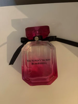 Victoria's Secret Bombshell parfym - Snygg parfymflaska från Victoria's Secret i modellen Bombshell. Flaskan är åttakantig med en rosa till transparent färgskiftning och har ett dekorativt svart band runt halsen. Perfekt för dig som gillar fräscha och feminina dofter.
