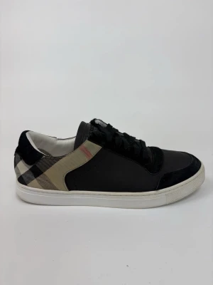 Burberry svart-beige låga sneakers - Snygga låga sneakers från Burberry i svart läder och mocka med klassiskt beige rutmönster på sidan. Skorna har vita sulor och svarta skosnören. Perfekt för dig som vill ha en stilren och tidlös look med en touch av lyx.