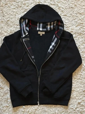 Svart hoodie med dragkedja Burberry - Svart hoodie från Burberry med klassiskt rutigt foder i huvan. Dragkedja framtill, fickor på sidorna och ribbade muddar. Perfekt för en stilren och avslappnad look.
