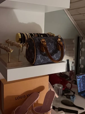  LVBLÅ denim handväska - Säljer en snygg blå denim handväska med klassiskt monogrammönster och detaljer i brunt läder. Väskan har två korta handtag och guldfärgade metalldetaljer. Perfekt för dig som vill ha en trendig och unik accessoar. Axelband medföljer