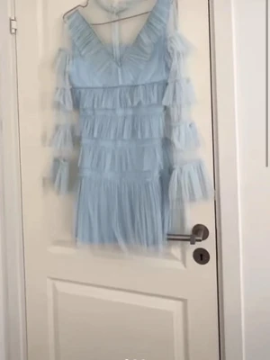 Ljusblå volangklänning med v-ringning - Supersöt ljusblå kortklänning med flera lager volanger och v-ringning. Klänningen har långa ärmar med volangdetaljer och är tillverkad i ett tunt, skirt material som ger en drömmig känsla. Perfekt för dig som vill sticka ut med en romantisk look.