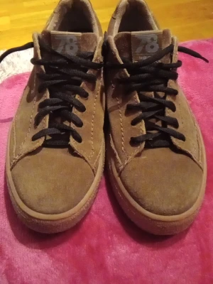 Bruna mockasneakers från 78 (BUGATTI) - Snygga bruna sneakers i mocka från märket 78. Skorna har svarta skosnören, låg profil och en klassisk siluett. Perfekta för dig som gillar en stilren och avslappnad look. Yttermaterialet är mjukt mockaläder och sulan är i gummi.