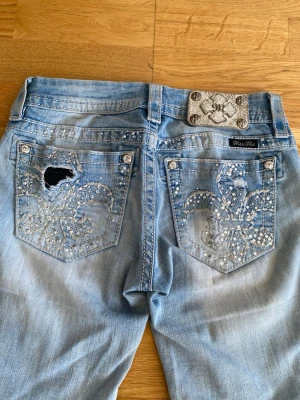 Ljusblå Miss Me jeans - Säljer ett par ljusblå jeans från Miss Me med stora bakfickor dekorerade med strass och broderade mönster. Jeansen har en design med slitning och hål på ena bakfickan samt en snygg metallpatch i midjan. Nypris runt 1600kr                      Kan posta samma dag🌸