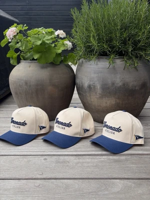 Voronado Riviera Cap - Snygg keps från Voronado Atelier i beige med blå skärm och broderad logga framtill. Klassisk sexpanel-design med justerbar rem bak. Perfekt accessoar för en avslappnad och trendig look.
