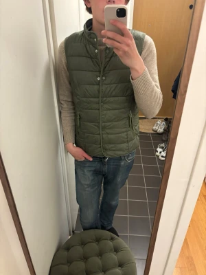 Olivgrön dunväst från Tom Tailor - Snygg olivgrön dunväst från Tom Tailor med ståkrage och dragkedja framtill. Västen har två sidofickor och quiltad design som ger en modern look. Perfekt att slänga över en hoodie eller tröja för extra värme. Materialet är lätt och smidigt. Finns en fläck på insidan