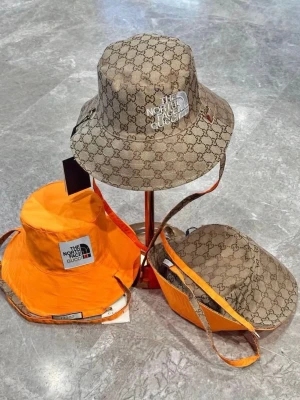 Bucket hat från The North Face x Gucci - Snygg bucket hat i samarbete mellan The North Face och Gucci. Hatten har Guccis klassiska monogrammönster i beige och brunt, samt detaljer i orange. Logga framtill och justerbart band. Perfekt accessoar för en trendig look.