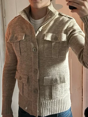 Beige Lambretta cardigan - Snygg beige stickad kofta med fyra stora fickor framtill och ribbad hög krage. Koftan har knäppning med stora knappar och dragkedja samt ribbade muddar vid ärmslut och nederkant. Perfekt som tröja på vintern eller jacka på våren!