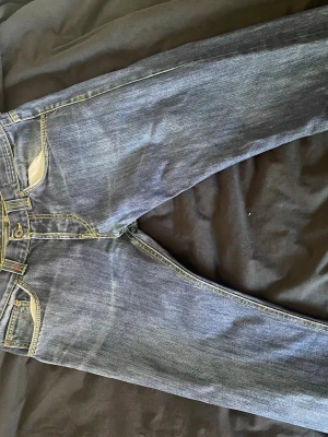 Mörkblåa jeans - Ett par jeans med raka ben och normalhög midja. Jeansen har en klassisk femficksmodell med två framfickor, en liten myntficka och två bakfickor. Gylfen stängs med dragkedja och knapp. De har kontrasterande sömmar i gult och orange, vilket ger en distinkt detalj till den mörkblå denimfärgen. Jeansen har bälteshällor runt midjan. Har tappat några knappar där bak, även lite sliten där nere men annars väldigt unika och snygga enligt kompisar. Kan fixa mått vid behov.