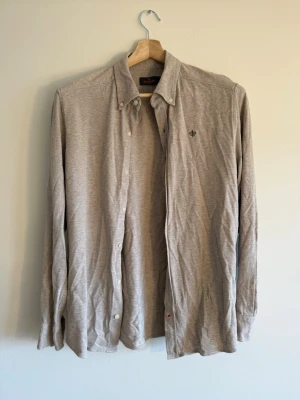 Morris Stockholm - Light Grey shirt - Morris Stockholm button-down i beige/grå bomullsjersey, stl M. Diskret fleur-de-lis-emblem. Neutral colorway som funkar under kavaj eller standalone. Skick: mycket bra