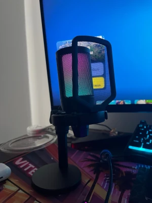 RGB USB-mikrofon med stativ - Snygg USB-mikrofon med RGB-belysning som skiftar färg. Mikrofonen har ett svart metallhölje och står på ett stabilt, runt stativ. Perfekt för gaming, streaming eller poddande. Enkel att koppla in via USB och passar snyggt på skrivbordet.