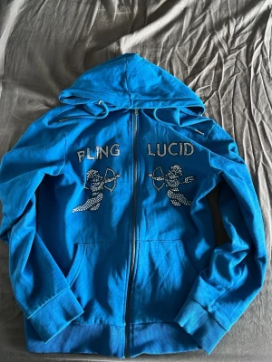 Blå hoodie från rhinestone med bling - Snygg blå hoodie från LAMODE med dragkedja och huva. Framsidan har blingade texter 'BLING' och 'LUCID' samt två änglar i strass. Mjuk insida och ribbade muddar. Perfekt för dig som gillar statement-plagg och vill sticka ut.
