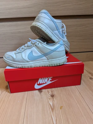 Nike Dunk Low i beige och ljusblå - Nike Dunk Low sneakers i beige med ljusblå detaljer och swoosh. Klassisk siluett med lågt skaft, ovandel i läder och meshfoder för extra komfort. Snygga och stilrena sneakers som passar till många outfits.