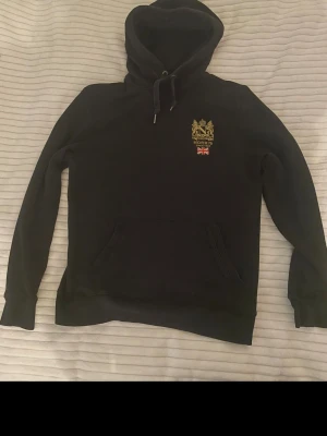 Morris hoodie - Svart hoodie från Morris Stockholm med broderad logga och brittisk flagga på bröstet. Klassisk känguruficka framtill och justerbar huva med dragsko. Tillverkad i mjukt bomullsmaterial, perfekt för en chill och stilren look.