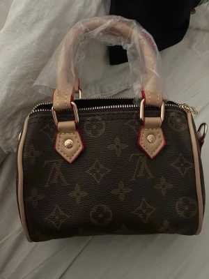  Monogram handväska - Snygg handväska i klassiskt brunt monogrammönster. Väskan har ljusa handtag med röda detaljer och guldfärgade metalldetaljer. Tillverkad i canvas med läderdetaljer och dragkedja upptill. Perfekt för dig som gillar lyxiga accessoarer.