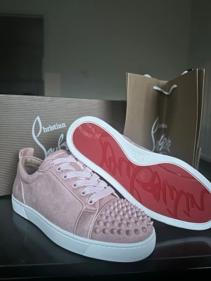 Rosa Christian Louboutin sneakers - Snygga rosa sneakers från Christian Louboutin i mocka med ikoniska nitar på tån och vit sula. Skorna har klassiska vita skosnören och den välkända röda sulan med logga. Perfekta för dig som vill sticka ut med exklusiv streetstyle.