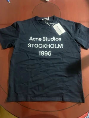 Marinblå Acne Studios t-shirt - Snygg marinblå t-shirt från Acne Studios med vit text 'Acne Studios STOCKHOLM 1996' på bröstet. Klassisk passform med rund hals och korta ärmar. Tillverkad i mjuk bomull, perfekt för en clean och stilren look.