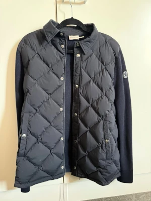 Moncler cardigan  - Cardigan från Moncler med quiltad front och stickade ärmar. Jackan har krage, tryckknappar och Moncler-logga på ärmen. Perfekt för dig som vill ha en stilren och exklusiv look. En liten defekt på dragkedjan. 