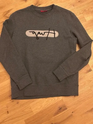 Hugo boss Mörkgrå sweatshirt - Mörkgrå sweatshirt med rund halsringning och svart/vitt grafiskt tryck på bröstet. Tröjan har långa ärmar, ribbade muddar och är tillverkad i mjukt bomullsmaterial. Perfekt för en avslappnad och stilren look.