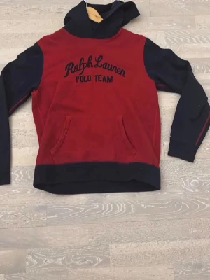 Röd och marinblå hoodie från Ralph Lauren - Snygg hoodie från Ralph Lauren Polo Team i rött och marinblått. Stor huva, magficka och broderad logga framtill. Perfekt för en avslappnad och sportig stil. Mjukt material som känns skönt mot huden.