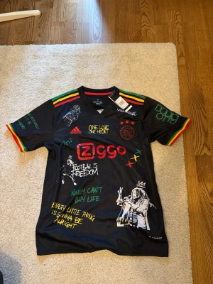 Ajax x Adidas svart fotbollströja L - Säljer en svart Ajax fotbollströja från Adidas limited edition Bob marley X Ajax med färgglada detaljer i rött, gult och grönt på axlarna. Tröjan har tryck med graffiti-inspirerade texter och motiv, samt Ziggo-loggan på bröstet. Materialet är lätt och andas, perfekt för träning eller match. 