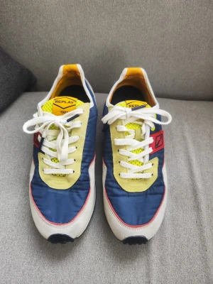 Retro färgglada herr sneakers från Replay  - Snygga sneakers från Replay i blått, gult och vitt med röda detaljer. Ovandelen är i textil och mocka, med vit sula och svarta samt orange detaljer på hälen. Klassisk låg modell med vita skosnören och mesh vid plösen. Finns tecken på användning. 