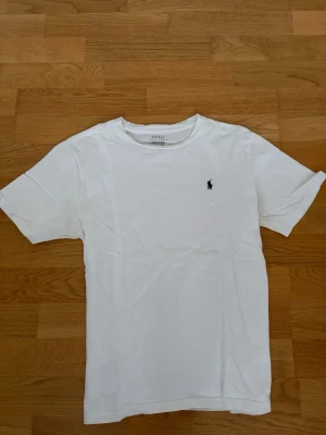 Vit t-shirt från Ralph Lauren - Klassisk vit t-shirt från Ralph Lauren med rund halsringning och korta ärmar. Smått missfärgad under armarna pga solkräm, lätt att ta bort själv. T-shirten har en liten broderad logga på bröstet och är tillverkad i mjuk bomull. Perfekt basplagg med stilren design. Storleken är L i barnstorlek 14-16 år men passar som S i herr och är slimfit. Hör gärna av dig vid frågor eller funderingar! Nypris runt 800 kr. 