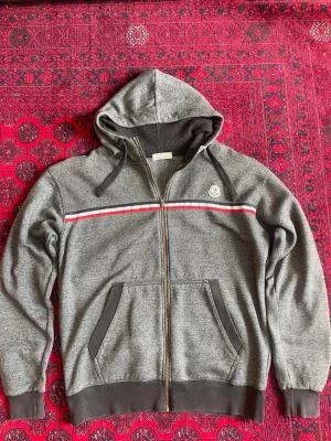 Grå Moncler hoodie med dragkedja - Snygg grå hoodie från Moncler med dragkedja, huva och fickor framtill. Dekorativt band i rött, vitt och blått över bröstet samt Moncler-logga på vänster sida. Muddar vid ärmslut och nederkant. Perfekt för en chill och sportig stil. Storlek L. 