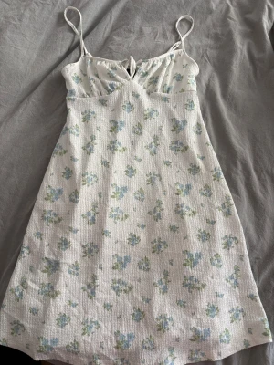 Vit blommig sommarklänning med snörning - Söt vit kortklänning från Gina tricot med blått och grönt blommönster. Klänningen har tunna axelband och snörning framtill vid bysten. Tillverkad i ett lätt, strukturerat bomullstyg – perfekt för varma dagar.
