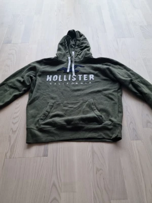 Olivgrön hoodie från Hollister - Snygg olivgrön hoodie från Hollister med vit logga och texten 'California' på bröstet. Hoodien har en stor känguruficka framtill, dragsko i huvan och är tillverkad i mjukt material. Perfekt för en avslappnad och trendig look.