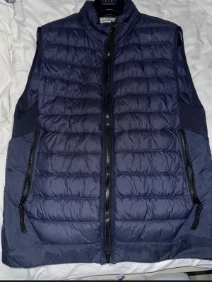 Mörkblå dunväst från Stone Island - Snygg mörkblå dunväst från Stone Island med quiltad design och dragkedja framtill. Västen har två praktiska fickor med dragkedja och den ikoniska Stone Island-loggan på sidan. Perfekt för lager-på-lager och streetwear-stil.