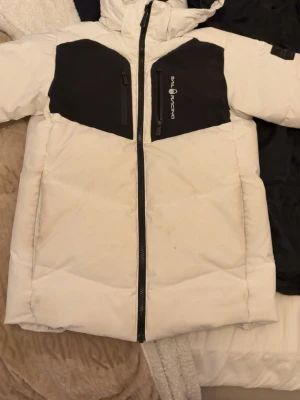 Vit pufferjacka från Sail Racing - Snygg vit pufferjacka från Sail Racing med svarta detaljer på bröstet och dragkedja framtill. Jackan har hög krage och huva, samt två bröstfickor med dragkedja. Perfekt för kalla vinterdagar och riktigt stilren look.