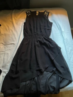 Svart klänning med paljetter KappAhl - Svart ärmlös klänning från KappAhl med snygga paljettdetaljer på axlarna. Klänningen har omlottliknande nederdel och markerad midja. Perfekt för dig som vill sticka ut lite extra med glittriga detaljer.