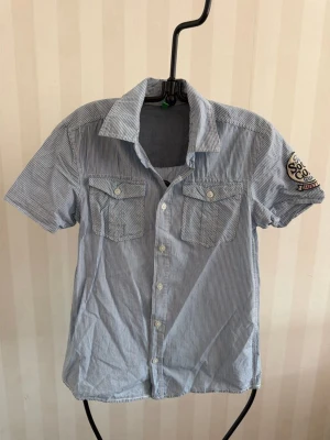 Blårandig kortärmad skjorta Benetton - Snygg blårandig skjorta från Benetton med korta ärmar, klassisk krage och två bröstfickor med knappar. Skjortan har ett broderat märke på ena ärmen och stängs med vita knappar framtill. Perfekt för en avslappnad stil.